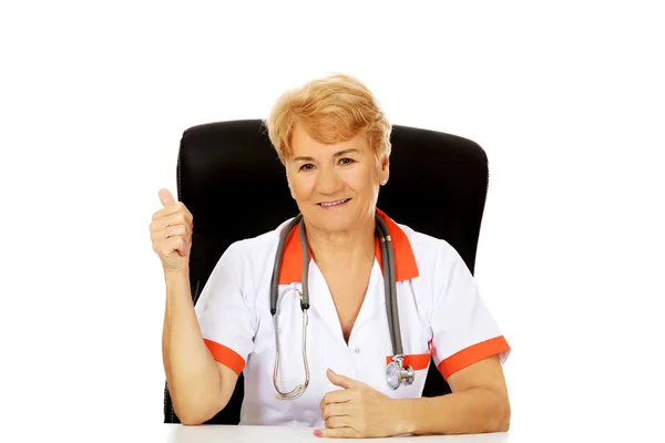 Sorria idosa médica ou enfermeira sentada atrás da mesa e mostra o polegar para cima — Fotografia de Stock