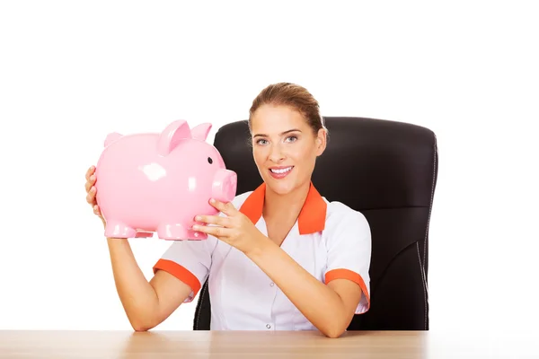 Smile doctor sitting behind the desk and holding a piggybank — Stock Photo, Image