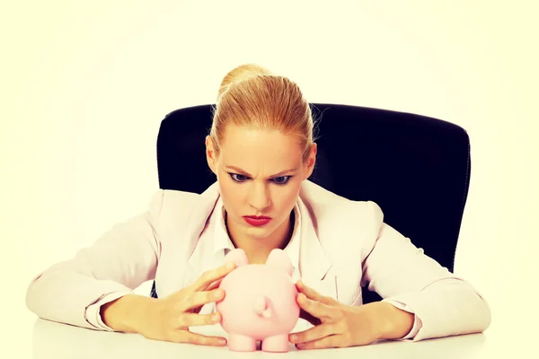 怒っているビジネスの女性彼女の piggybank を探していると、机の後ろに座っています。 — ストック写真