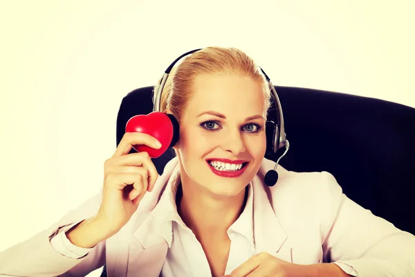 Happy call center woman sitting behind the desk and holding heart model — Stock Photo, Image