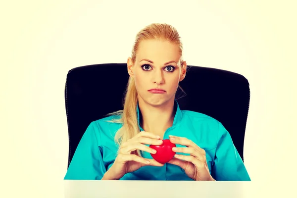 Médica ou enfermeira preocupada sentada atrás da mesa e segurando modelo de coração — Fotografia de Stock