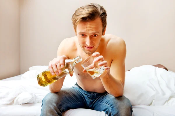 Пьяный мужчина сидит на кровати и пьет виски — стоковое фото
