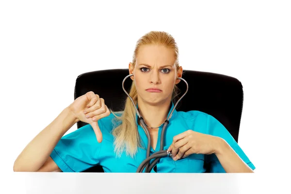 Nieszczęśliwa kobieta lekarz lub pielęgniarka siedzi za biurkiem z stetoskop i pokazuje kciuk w dół — Zdjęcie stockowe