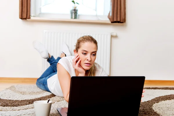 Yerde yatıp kahve içerken dizüstü bilgisayar kullanan odaklanmış genç bir kadın. — Stok fotoğraf