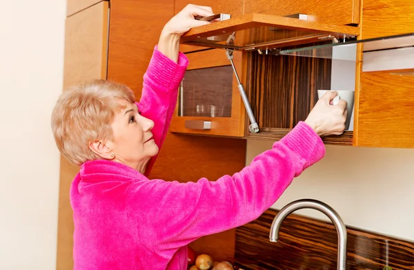 Leende senior kvinna tar mugg från ett köksskåp — Stockfoto