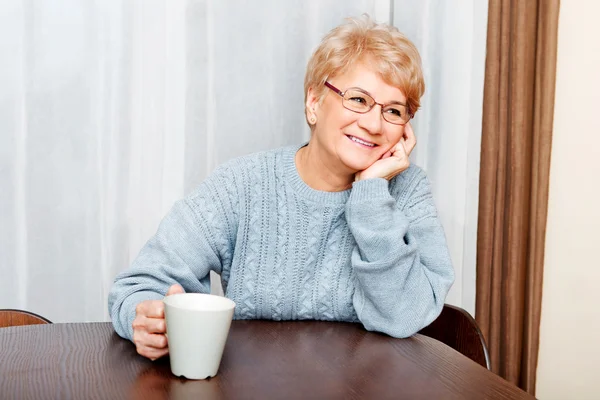 Senior woman sitting at the desk and drinking coffee or tea — Stock Photo, Image