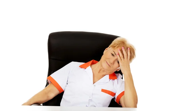 Médica idosa ou enfermeira sentada atrás da mesa com dor de cabeça — Fotografia de Stock