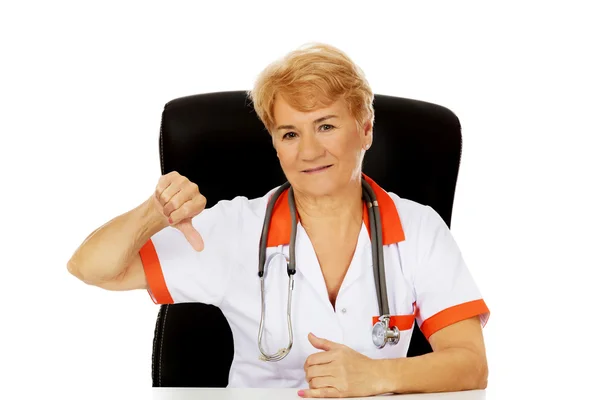 Médico o enfermera anciana sentada detrás del escritorio y muestra el pulgar hacia abajo —  Fotos de Stock