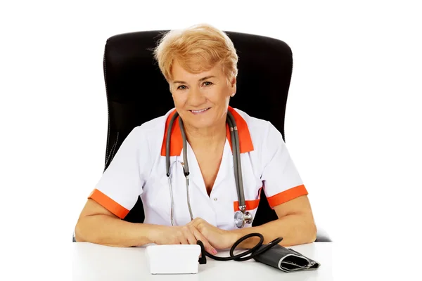 Smile elderly female doctor or nurse sitting behind the desk with bloog preasure gauge — Stock Photo, Image