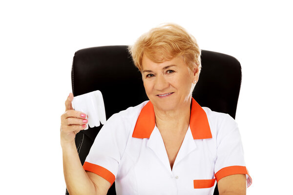 Smile elderly female dentist sitting behind the desk and holds big tooth model