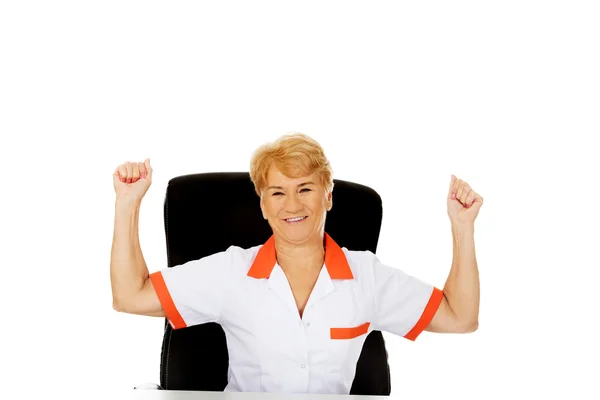 Gelukkig oudere vrouwelijke arts of verpleegkundige vergadering achter het Bureau withd handen omhoog — Stockfoto