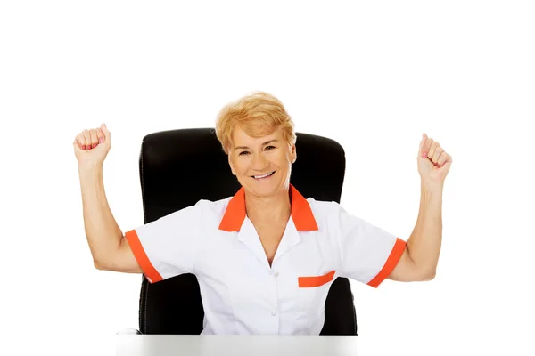 Happy elderly female doctor or nurse sitting behind the desk withd hands up — Stock Photo, Image