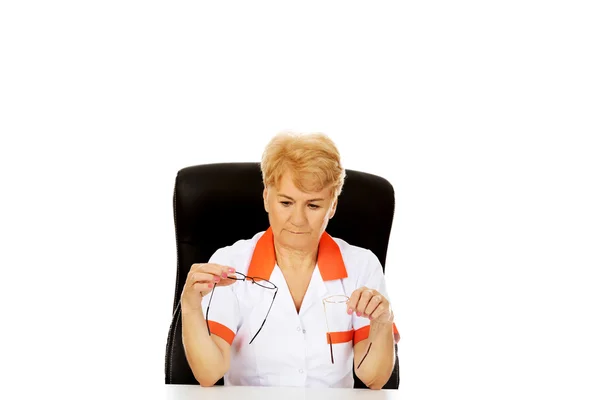 Worried elderly female doctor or nurse sitting behind the desk and holds two pair of glasses — Stock Photo, Image