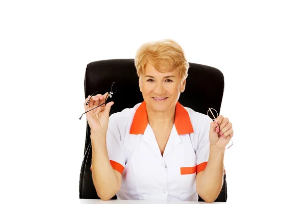 Sorria idosa médica ou enfermeira sentada atrás da mesa e segura dois pares de óculos — Fotografia de Stock