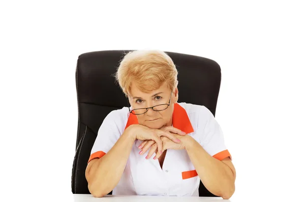 Worried elderly female doctor or nurse sitting behind the desk — Stock Photo, Image
