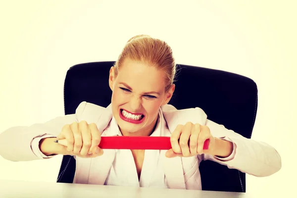 Angry business woman sitting behind the desk and trying to break huge pen — Stock Photo, Image