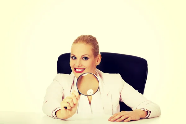Mujer de negocios sonrisa sentado detrás del escritorio y mirando en una lupa — Foto de Stock