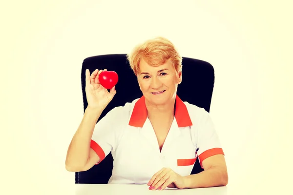Uśmiech starszych kobiet lekarz lub pielęgniarka siedzi za biurkiem i posiada serce zabawka — Zdjęcie stockowe