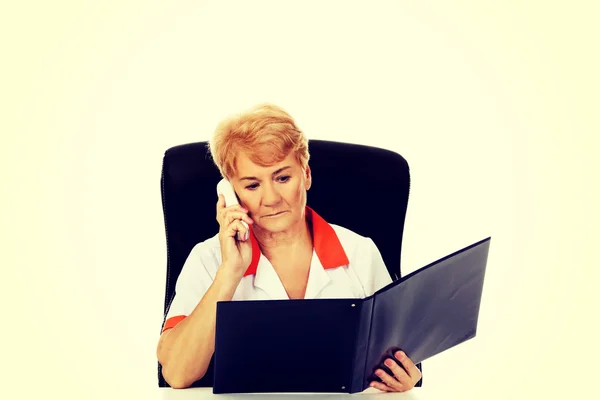Worried elderly female doctor or nurse sitting behind the desk and talking through a phone — Stock Photo, Image