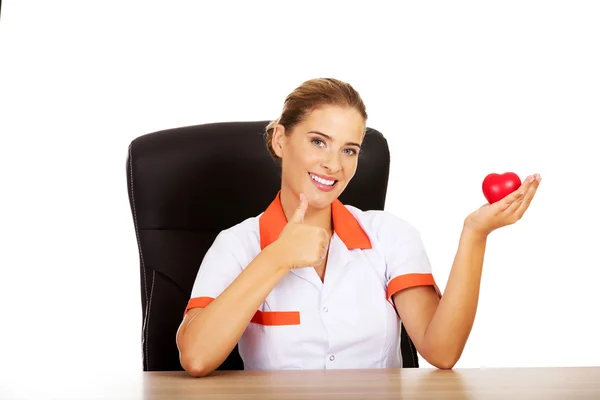 Ärztin sitzt hinter dem Schreibtisch und hält Herzspielzeug — Stockfoto