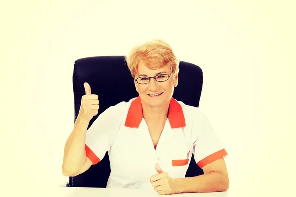微笑老年女性医生或护士坐在桌子和节目拇指后面 — 图库照片