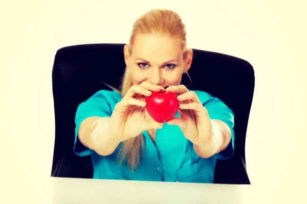 Médico o enfermera sonriente sentada detrás del escritorio y sosteniendo el juguete del corazón —  Fotos de Stock