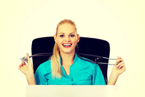 Забавная женщина-врач или медсестра сидит за столом со стетоскопом — стоковое фото