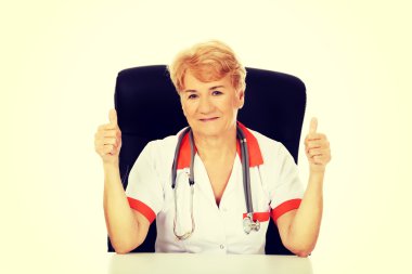 Yaşlı kadın doktor veya hemşire Resepsiyon ve gösterileri başparmak oturan gülümse