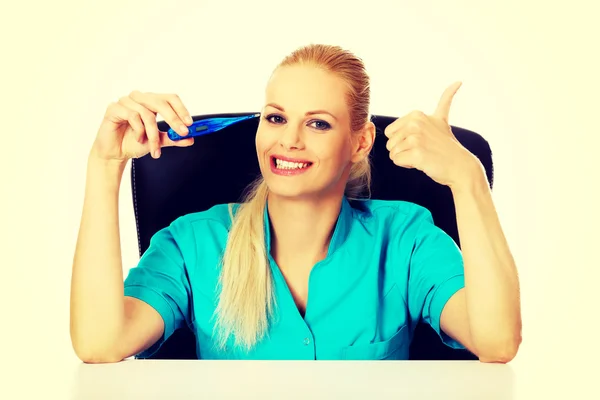 Médico o enfermera sonriente sentada detrás del escritorio sosteniendo el termómetro y mostrando el pulgar hacia arriba —  Fotos de Stock