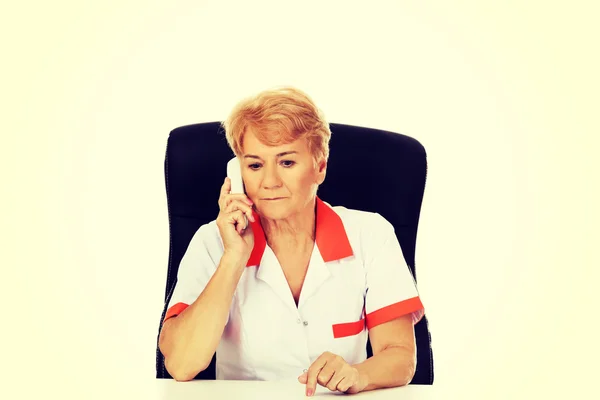 Endişeli yaşlı kadın doktor veya hemşire masanın arkasında oturan ve bir telefondan konuşuyor — Stok fotoğraf