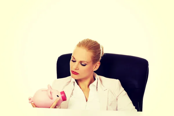 Wütende Geschäftsfrau sucht ihr Sparschwein und sitzt hinter dem Schreibtisch — Stockfoto