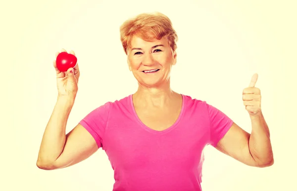 Old smiling woman holding red toy heart with thumb up — Stock Photo, Image