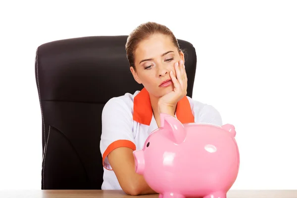Female doctor sitting behind the desk and holding a piggybank Stock Picture