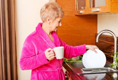 Son sınıf öğrencisi kadın mutfakta kahve içiyor ve tabak yıkıyor.