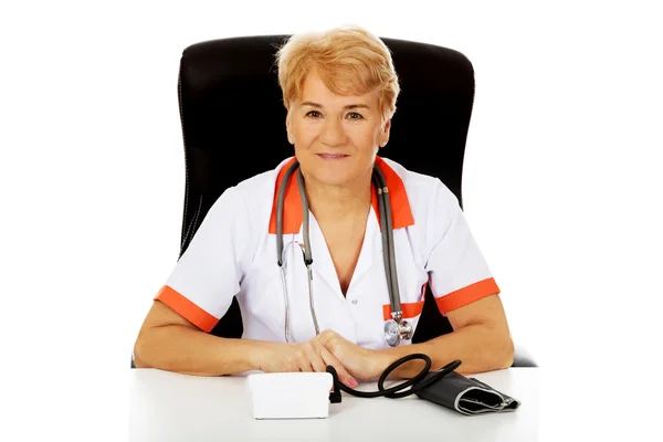 Smile äldre kvinnliga läkare eller sjuksköterska sitter bakom skrivbordet med bloog preasure mätare — Stockfoto