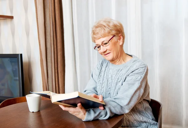 Starší žena sedí u stolu a číst knihu a pití čaje — Stock fotografie