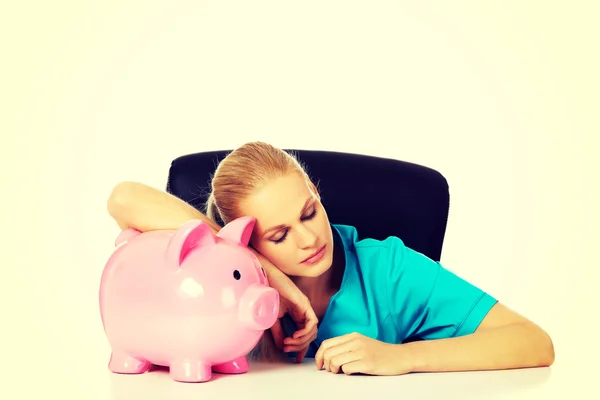 Junge müde Ärztin oder Krankenschwester, die hinter dem Schreibtisch sitzt und ein Sparschwein hält — Stockfoto