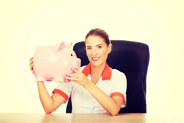 Leende läkare sitter bakom skrivbordet och hålla en piggybank — Stockfoto