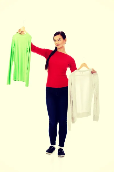 Smiling hesitant young woman holding two shirts — Stock Photo, Image