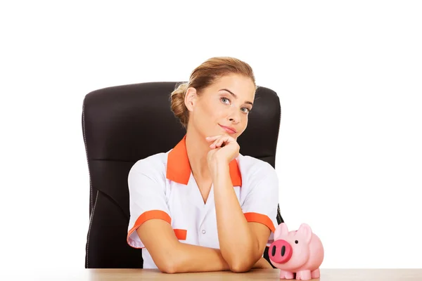 Female doctor sitting behind the desk and holding a piggybank — Stock Photo, Image