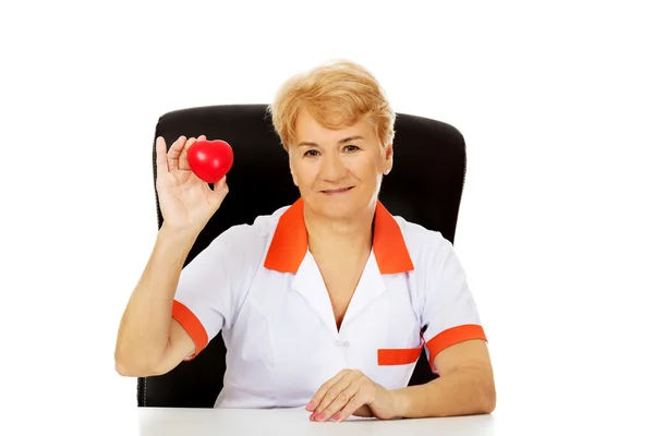 Glimlach van oudere vrouwelijke arts of verpleegkundige vergadering achter de balie en houdt hart speelgoed — Stockfoto