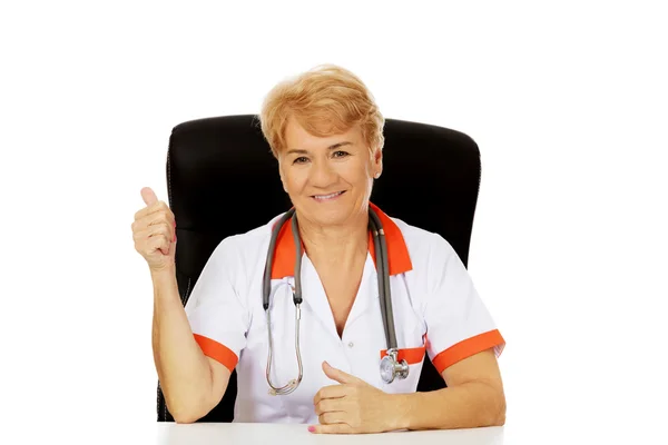 Улыбайтесь, пожилая женщина врач или медсестра сидит за столом и показывает большой палец вверх — стоковое фото
