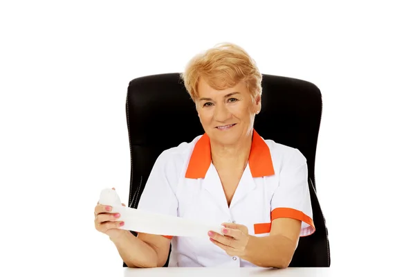 Souriez femme âgée médecin ou infirmière assis derrière le bureau et tient bandage — Photo