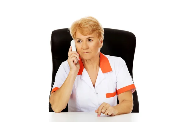 Worried elderly female doctor or nurse sitting behind the desk and talking through a phone — Stock Photo, Image