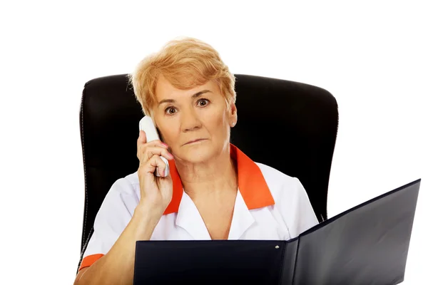 Worried elderly female doctor or nurse sitting behind the desk and talking through a phone — Stock Photo, Image