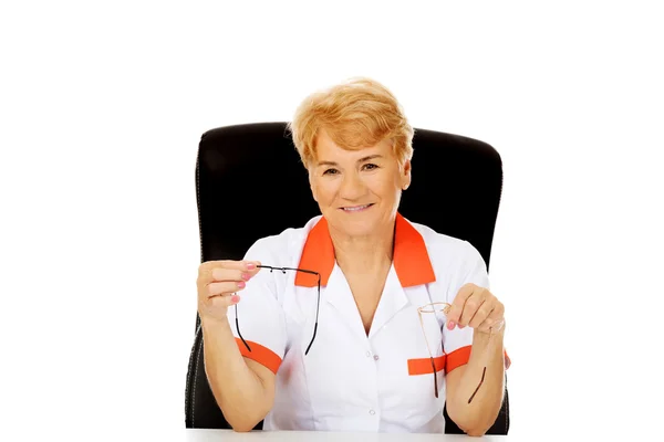 Smile elderly female doctor or nurse sitting behind the desk and holds two pair of glasses — Stock Photo, Image