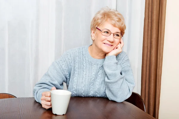 Senior woman sitting at the desk and drinking coffee or tea — Stock Photo, Image