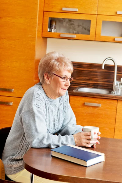 Старша жінка сидить за столом з книгою і п'є чай або каву — стокове фото