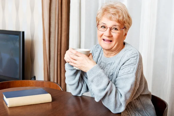 年配の女性の本と机に座って、お茶やコーヒーを飲む — ストック写真