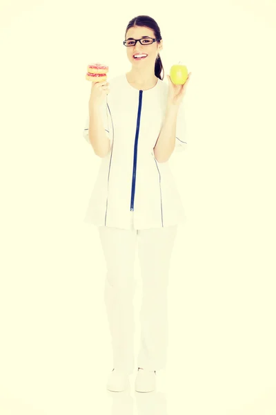 Young female doctor, nurse holding an apple and doughtnuts. — Stock Photo, Image